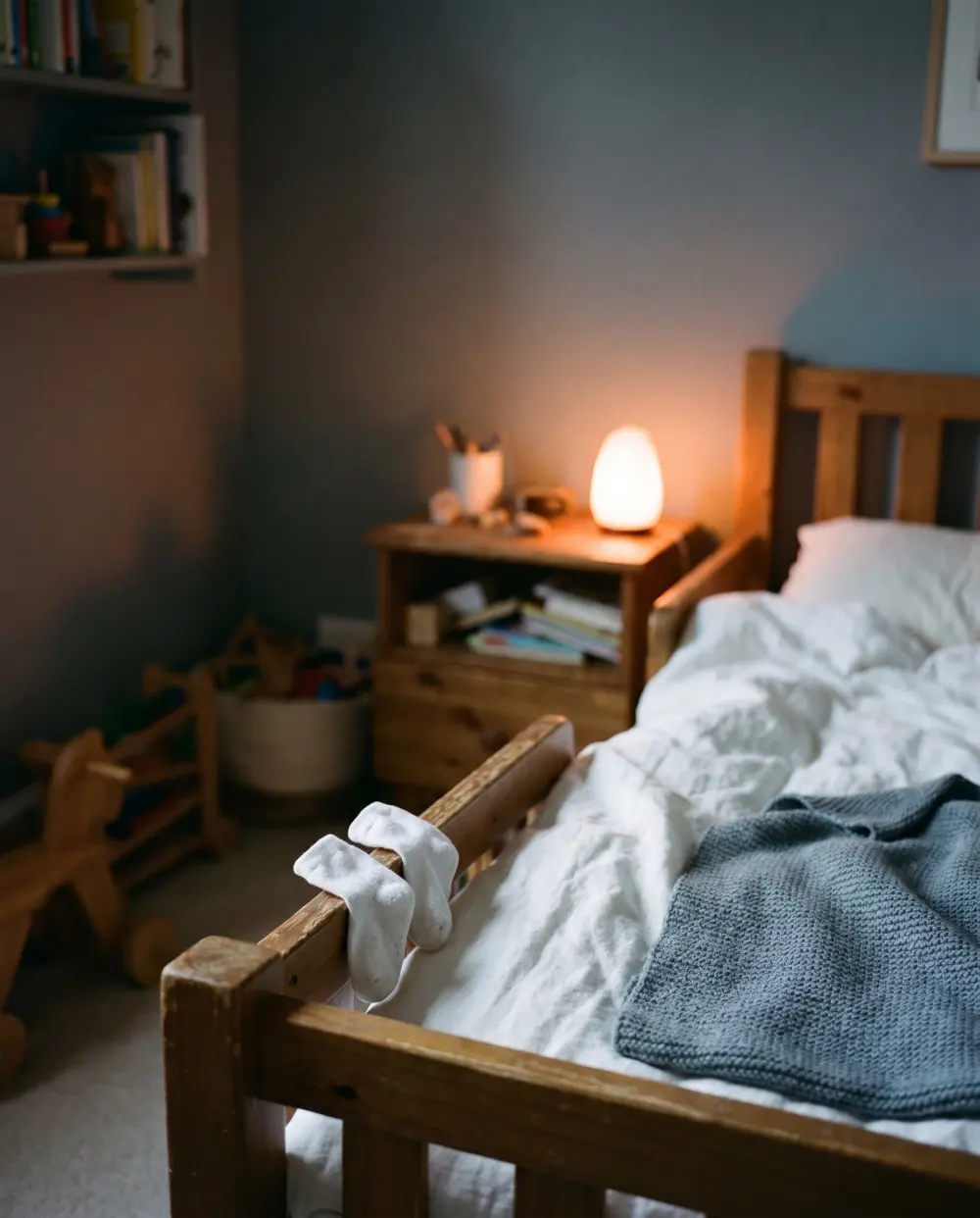 Weiße Kindersocken hängen nachts am Gitter eines Kinderbetts, daneben brennt eine Nachttischlampe